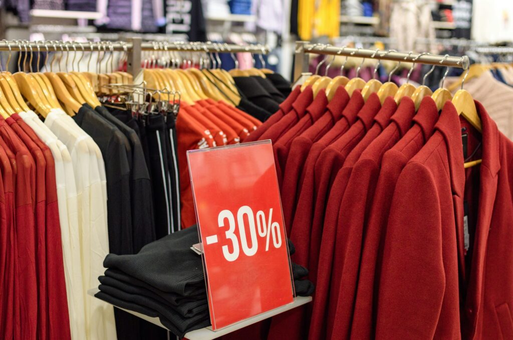 Red discount sign in retail store with clothes on coat hangers.