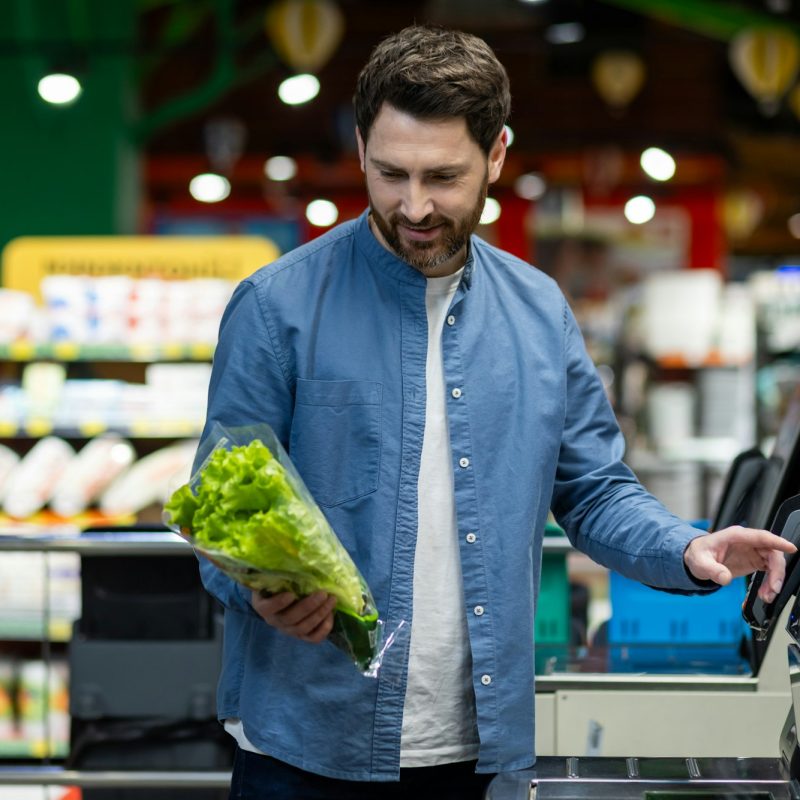 Man using self-checkout at grocery store while holding fresh lettuce, exemplifying modern shopping