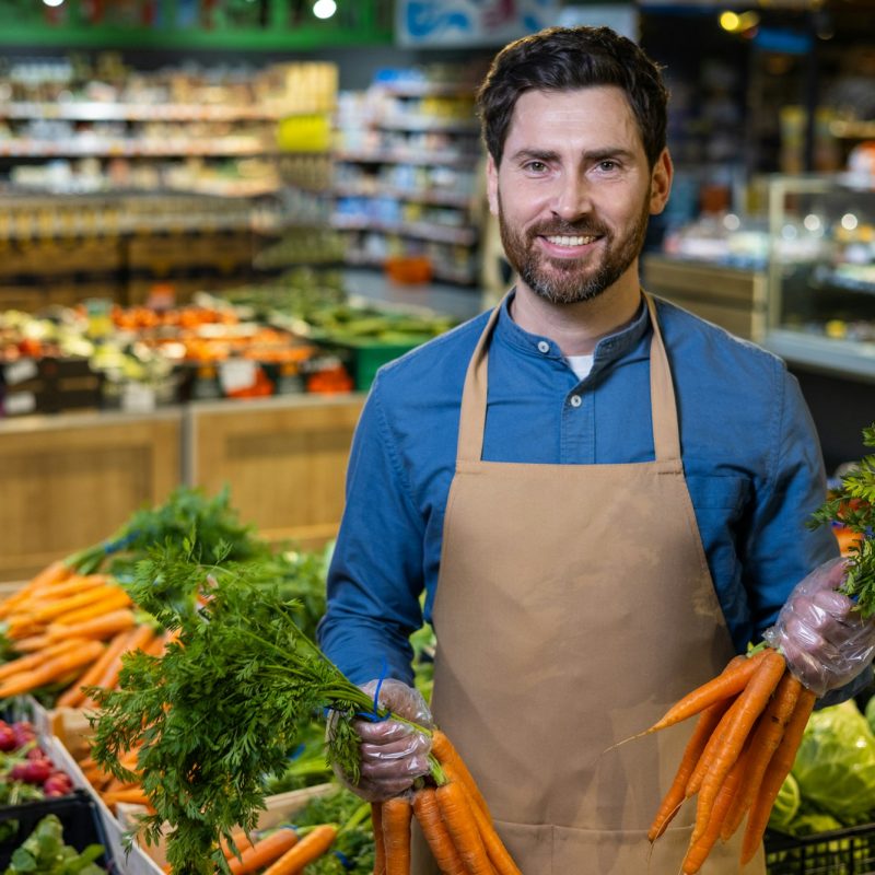 Smiling grocery store worker holding fresh carrots in vibrant vegetable aisle of supermarket